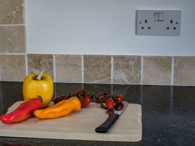 A wooden chopping board on a kitchen counter with a yellow bell pepper red and orange peppers cherry tomatoes on the vine and a knife at The Folly in Port Isaac