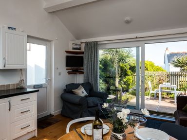 A kitchen and living area with a dining table set with plates and wine a chair a door a TV and garden view at The Folly in Port Isaac