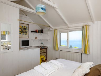 A bedroom with a bed folded towels a small tv shelves a window with striped curtains and an ocean view at The Folly in Port Isaac
