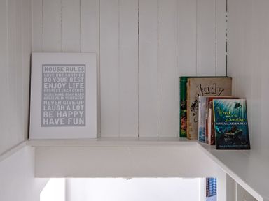 A white wooden shelf with a framed house rules print and a row of books on a white paneled wall at The Folly in Port Isaac