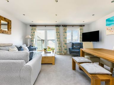 A living room with grey sofas a wooden coffee table a blue armchair a wooden dining table with cushioned benches and chairs a wall mounted tv and windows with floral curtains at Rock Lobster in Port Isaac