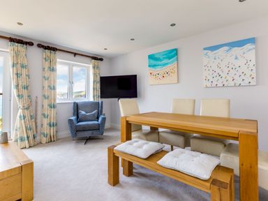 A dining area with a wooden table, white chairs, a wooden bench with cushions, a blue armchair, curtains with blue patterns, a wall-mounted TV, and two paintings on the wall at Rock Lobster in Port Isaac