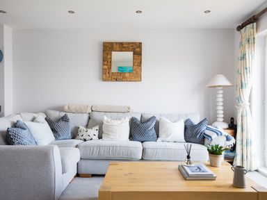 A living room with a gray sectional sofa with pillows a wooden coffee table and a lamp near a window at Rock Lobster in Port Isaac