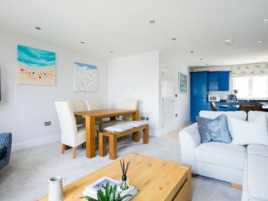 A dining area with a wooden table and white chairs next to a gray sofa and blue kitchen cabinets at Rock Lobster in Port Isaac