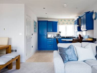 Open plan living area with blue kitchen cabinets wooden dining table and light gray sofa with blue cushions