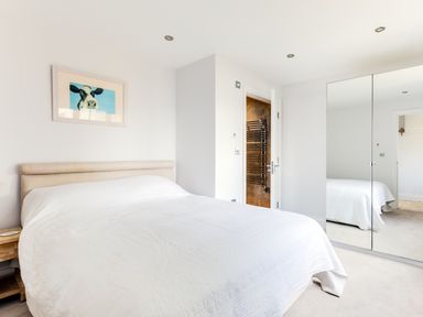 A bedroom with a beige headboard bed covered in a white bedspread a wooden bedside table a framed picture of a cow a door leading to a bathroom and mirrored wardrobes at Rock Lobster in Port Isaac