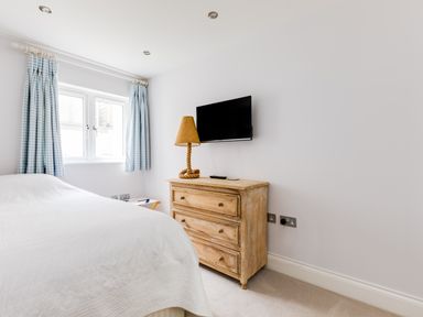 A bedroom with a bed white bedspread wooden chest of drawers table lamp and television on the wall at Rock Lobster in Port Isaac