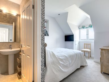 A bedroom with a bed covered in white bedding a wooden chair a wall-mounted television a wooden dresser and an open door leading to a bathroom with a sink and mirror at Rock Lobster Port Isaac