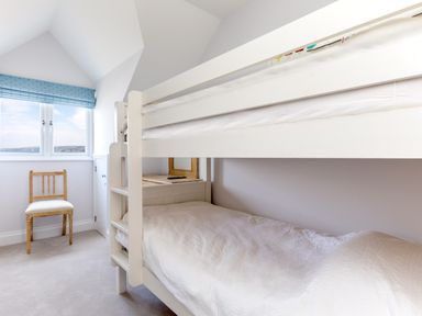 A bedroom with white bunk beds a wooden chair near a window with blue blinds and a side table at Rock Lobster in Port Isaac