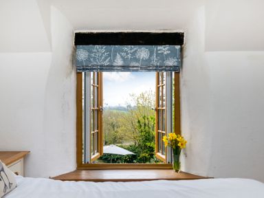 A window with floral curtains and a vase of yellow flowers on the sill overlooking trees and an umbrella at Dingles Cottage in Rock