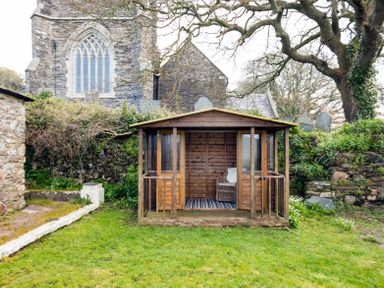 A small wooden garden shed with a chair and rug inside in a grassy yard near a stone building and trees at Dingles Cottage in Rock