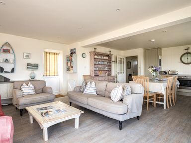 An open living and dining area with beige sofas, a wooden coffee table, a dining table with chairs, a kitchen area, and a wooden cabinet at Atlantis in Polzeath