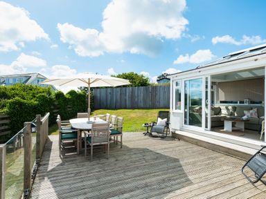A wooden deck with a dining table and chairs under a large umbrella next to a garden and a room with a sofa at Atlantis in Polzeath