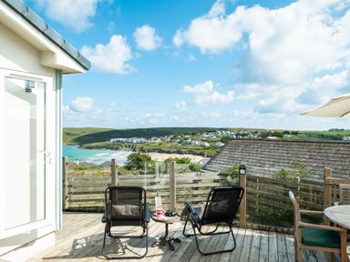 A wooden deck with two chairs a small table and a view of a beach and houses at Atlantis in Polzeath