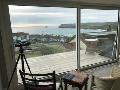 A room with a telescope, chairs, a small wooden table, and a glass door overlooking a deck and coastal houses at Atlantis Polzeath