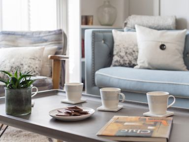 A coffee table with three cups on coasters a plate of biscuits a plant and a book in a living room with blue and patterned chairs at Albany in Port Isaac