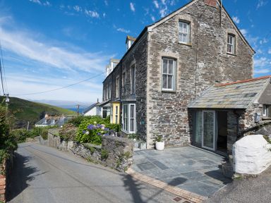 A stone house with a slate patio and flowers next to a narrow road with hills in the background at Albany in Port Isaac