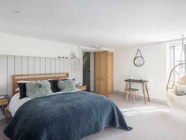 A bedroom with a bed covered with a dark blue quilt and green pillows a wooden door open a table with a mirror and a hanging chair at Albany in Port Isaac