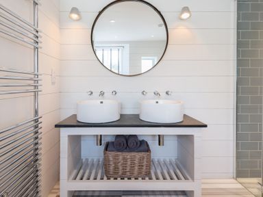 A bathroom with two round white sinks on a black countertop a large round mirror above and a basket with towels below at Albany in Port Isaac