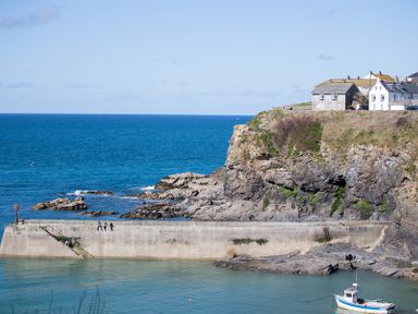 A sea harbor with a concrete pier and people standing on it near rocky cliffs with houses above and a small boat in the water at Albany in Port Isaac