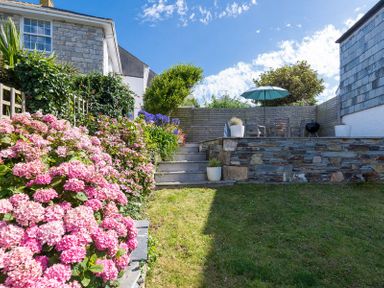 A garden with a lawn pink flowers stone steps and a patio with table chairs and an umbrella at Albany in Port Isaac