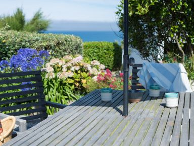 An outdoor wooden table with ceramic mugs and chairs with a garden and sea in the background at Albany in Port Isaac