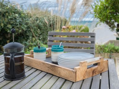 A wooden tray with three cups and stacked plates and a French press on a wooden outdoor table with a bench and plants at Albany in Port Isaac
