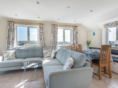 A living room with a light blue sectional sofa floral curtains and a dining table with wooden chairs at Polmorla in Polzeath