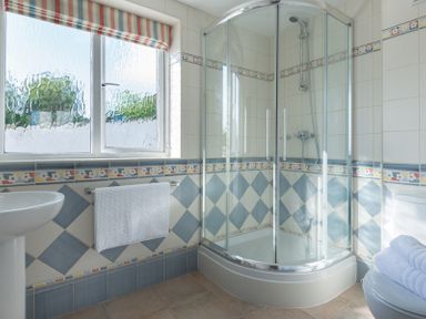 A bathroom with a corner shower enclosure a white sink a toilet with towels and a window with a striped blind at Polmorla in Polzeath