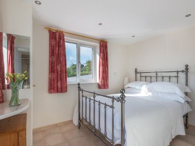 A bedroom with a metal bed frame white bedding red curtains and a vase with yellow flowers on a dresser at Polmorla in Polzeath