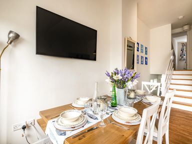 A dining area with a wooden table set for six with plates glasses and flowers on a table near a staircase and wall mounted TV at Slipway 12 in Rock