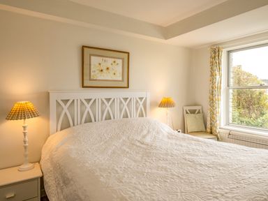 A bedroom with a white bed frame bedside tables lamps a chair and a window with floral curtains at Slipway 12 in Rock