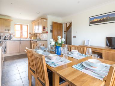 A dining table set with bowls glasses and cutlery next to a kitchen with wooden cabinets at Lundy Cottage in Rock
