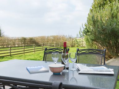 An outdoor table with a wine bottle three wine glasses a bowl a book and a newspaper with chairs and a fenced grassy area in the background at Lundy Cottage in Rock