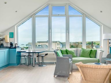A living room with a large window sofa with green cushions and kitchen area at The Point in Polzeath