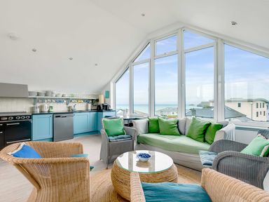A living room with wicker chairs a sofa with green cushions a round table and a kitchen area with blue cabinets at The Point in Polzeath