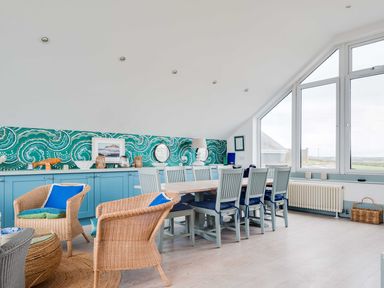 A dining area with a long table and multiple chairs near large windows and wicker armchairs at The Point in Polzeath