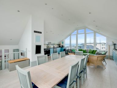 An open plan kitchen and dining area with a long wooden table and chairs a sitting area with green cushions and large windows showing a coastal view at The Point in Polzeath