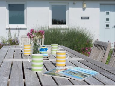 A wooden outdoor table with striped mugs a teapot a glass vase with flowers and magazines in a garden at The Point in Polzeath