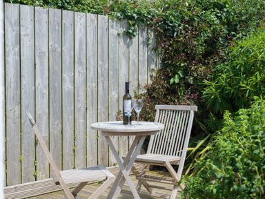 A wooden patio table with two wooden chairs a wine bottle and two wine glasses on the table with a wooden fence and plants in the background at The Point in Polzeath