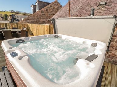 An outdoor hot tub with bubbling water on a wooden deck with stone buildings in the background at Cheviot Hills in Wooler