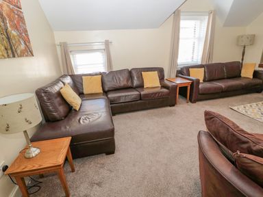 A living room with brown leather sofas and yellow cushions two wooden side tables with lamps and windows with beige curtains at Cheviot Hills in Wooler