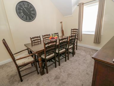 A dining room with a wooden table and eight chairs a fruit bowl on the table a window with curtains a large wall clock and a giraffe sculpture at Cheviot Hills in Wooler