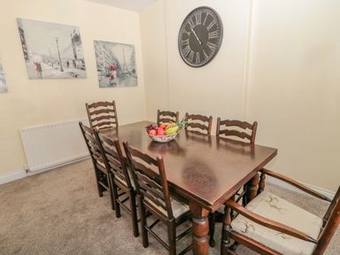 A dining room with a wooden table and six chairs a bowl of fruit on the table a large clock on the wall and two paintings on the wall at Cheviot Hills in Wooler