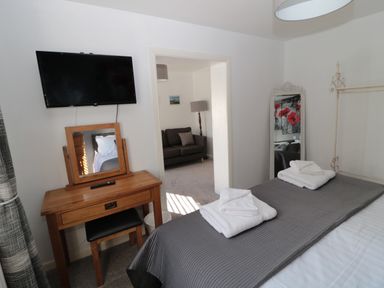 A bedroom with a bed, folded towels, a wooden dressing table with a mirror, a wall-mounted TV, and a doorway leading to a sitting room with a sofa at Cheviot Hills in Wooler