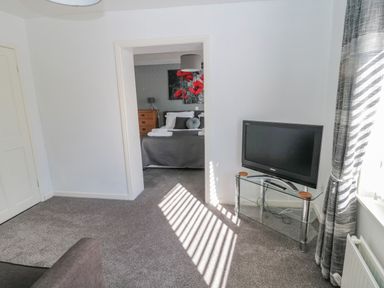 A room with a television on a glass stand and a view into a bedroom with a bed and artwork at Cheviot Hills in Wooler