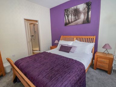 A bedroom with a wooden bed dressed in white and purple bedding two wooden nightstands with lamps and an open door showing a shower at Cheviot Hills in Wooler