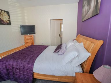 A bedroom with a wooden bed frame purple bedding a wooden chest of drawers a wall-mounted TV and a door at Cheviot Hills in Wooler