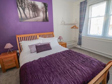 A bedroom with a wooden bed with purple bedding two bedside tables with lamps a garment rack and a window with curtains at Cheviot Hills in Wooler