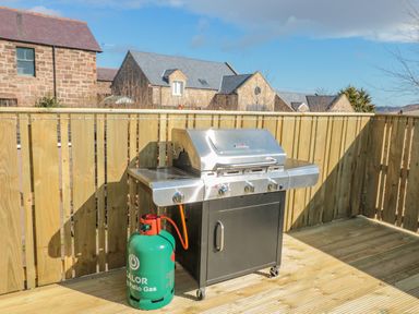 A gas grill with propane tank on a wooden deck with wooden fence and stone houses in the background at Cheviot Hills in Wooler